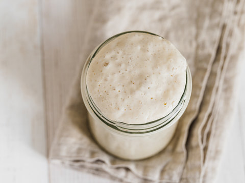 Wheat sourdough starter. Top view of glass jar with sourdough starter on white wooden background. Copy space for text or design.