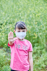 Niña con mascarilla en un ambiente ajardinado, con la palma extendida en señal de stop.
