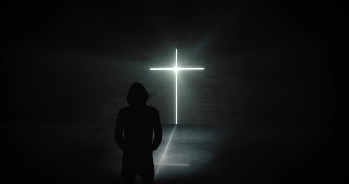 Mysterious person stands in a dark room in front of a bright glowing cross symbol