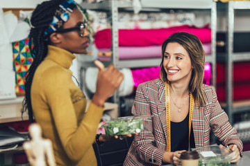Two young entrepreneur women, or fashion designers working in atelier and making short breakfast break at work