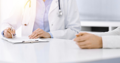 Unknown woman-doctor and female patient sitting and talking at medical examination in clinic, close-up. Therapist wearing blue blouse is filling up medication history record. Medicine concept