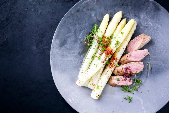 Fried Iberian pork fillet sliced with blanched white asparagus and herbs with spice as closeup on a modern design plate with copy space left