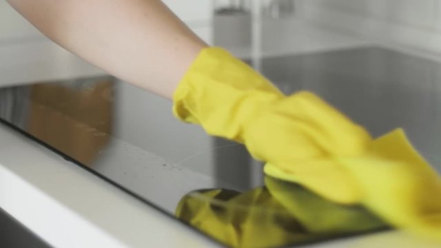 Cleaning cooktop cooking panel in kitchen with fat remover spray and yellow rag by a woman in yellow rubber gloves.
