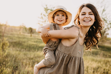 Young woman and little boy playing in sun light, outdoors, in a field. Mother gives a piggyback riding to her son, smiling, both wearing vintage clothes and straw hats.