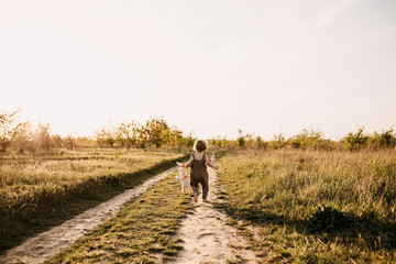 Little blonde boy with curly hair, wearing vintage jumpsuit, running on a dirt path, barefoot, at sunset, holding a plush rabbit toy.