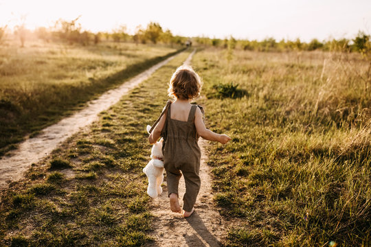Little blonde boy with curly hair, wearing vintage jumpsuit, walking on a dirt path, barefoot, at sunset, holding a plush rabbit toy.