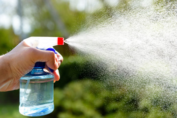 a man's hand sprays liquid from a spray on a background of greenery in the sun
