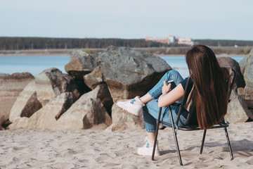 A long-haired brunette girl in blue jeans is sitting on a chair on the beach with a phone. Rear view. Sunny summer day
