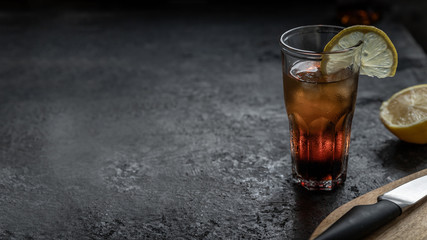 A glass of rum with cola full of ice with a slice of lemon on a dark gray concrete bar table. Big copy space
