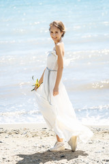 Fototapeta na wymiar Bride at the sea. Wedding ceremony on the sea, at the beach. Background of sea and mimosa tree . Young bride is dressed in white dress in greek style.