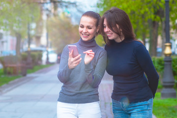 Happy Two girlfriends women looking at the phone and laughing. Walk in the park, girls look in a mobile smartphone