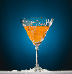 Frosted martini glass with orange liquid against a dark blue background and white floor