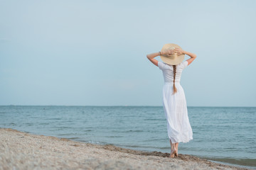 Fototapeta na wymiar Delicate young woman in white dress and hat walks on sea shore. Back view