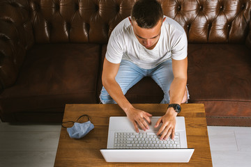 A young man working at a computer at home face mask lying on tables, working remotely during quarantine, pandemic COVID-19. Avoid the spread of coronavirus. Pandemic