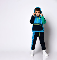 Little child in colorful sport suit and sneakers. He has put his hood on while posing isolated on white studio background