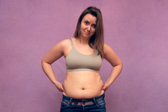 Overweight woman hand pinching excessive belly fat on pink background, unhealthy concept