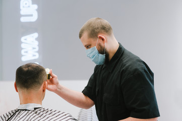 Male hairdresser in a medical mask cuts the client's hair with a clipper during the quarantine period. The work of a hairdresser in the quarantine of the coronavurus.