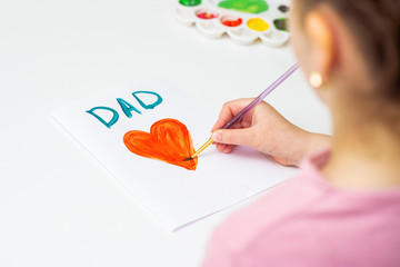 Child is drawing red heart with word Dad greeting card on white paper. Happy Father's Day concept.