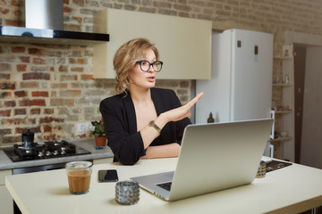 A woman in her kitchen works remotely on a laptop. A blond girl in glasses gesturing while talking to her colleagues in a video call at home.