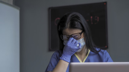 Exhausted doctor working with laptop computer. Tired woman rubs the nose to relieve tension from overtime work. Female medic resting from excessive workload. Overworking concept