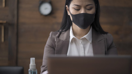 Asian woman in mask working computer in office with wooden wall on background. Beautiful woman wearing protective face mask works laptop. Pandemic concept. Front view