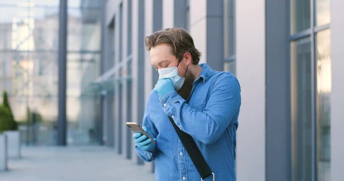 Male pedestrian in medical mask walking the street, caughing and texting message on mobile phone. Ill man in respiratory protection strolling outdoors and tapping or scrolling on smartphone.
