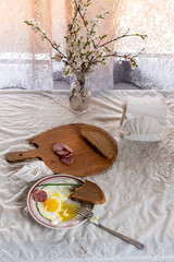 Fototapeta na wymiar Breakfast. Fried eggs of two eggs and green feathers of onions. Sausage and bread on a wooden board. Sprigs of blossoming apple tree in a crystal vase. Crumpled napkin.