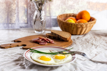 Breakfast. Fried eggs of two eggs and green feathers of onions. Sausage and bread on a wooden board. Sprigs of blossoming apple tree in a crystal vase. Apples and oranges in a wicker basket.