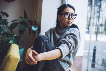 Contemplating Asian woman sitting on windowsill at home