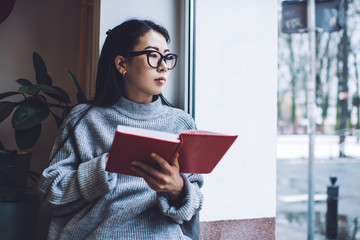 Pensive Asian woman reading notes in diary on windowsill