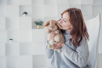 A young woman in a gray sweater is sitting at home in isolation with a rabbit on a light background