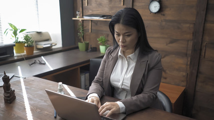 Asian businesswoman is typing on a computer taking off her mask while sitting alone at an office desk. Beautiful brunette working on a laptop in an empty office during quarantine