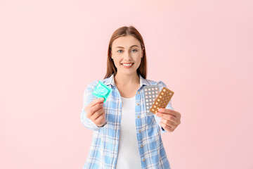 Young woman with condom and birth control pills on color background