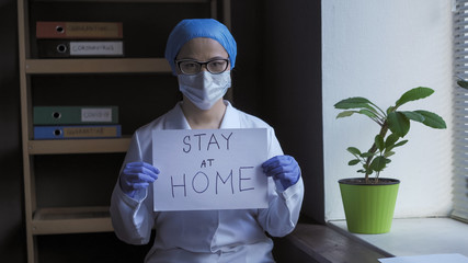 Stay at home slogan from tired doctor. Pandemic of Coronavirus concept. Female medic stands in medical office holding white paper blank