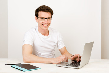 A handsome man with a light smile wearing a T-shirt and glasses sits against a white wall at his desk and work with a laptop. The concept of remote quarantine education and communication