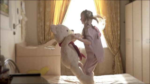 A girl with a polar bear jumps high in a large room. Day in on against a bright window
