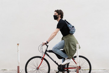 Young adult cyclist man walking in the city riding his bicycle. Hipster guy wearing a black safety mask over a bike against a white wall. Urban shot during coronavirus pandemic epidemics.