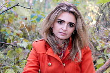 beautiful confident blond thoughtful woman with long hair in orange coat looking at camera and sitting in bushes in forest