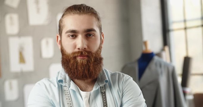Close-up portrait shot of bearded hipster clothing designer working in his office, looking at camera and positively smiling - small business, fashion concept 4k footage