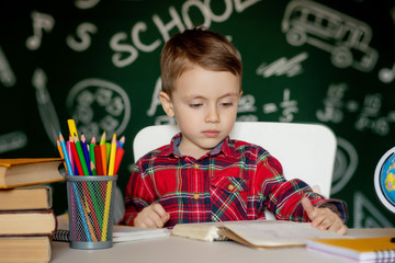 Cute clever boy is sitting at a desk with magnifying glass in hand. Child is reading a book with a blackboard on a background. Ready for school. Back to school