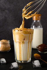 Closeup of Iced Dalgona coffee in glass mug and teaspoon on dark background. Trendy refreshment creamy whipped iced coffee. Korean coffee drink. Copy space