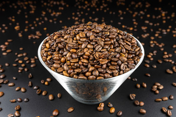 Fresh, roasted coffee beans in a glass bowl, standing on a black table, with lots of coffee scattered around.