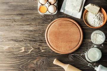 Baking cooking ingredients flour, eggs, rolling pin, butter, cottage cheese and a wooden round board on a wooden background. View from above. Copy space. Cookie pie or cake recipe layout