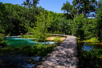 Wooden path built over the streaming waters of Krka National Park in Croatia - Tourists hiking in the forest along the river down to the waterfall in summer