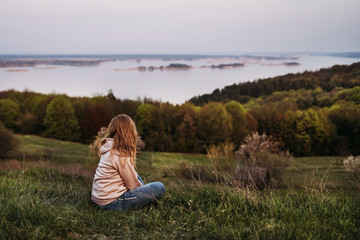 Fototapeta na wymiar A girl with blond hair sits on the grass and looks into the distance in a pink raglan with a hood. Sunset. Time for reflection. Inner peace. Psychological relaxation