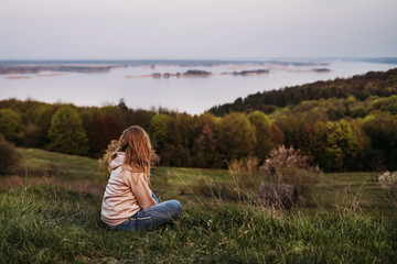  A girl with blond hair sits on the grass and looks into the distance in a pink raglan with a hood. Sunset. Time for reflection. Inner peace. Psychological relaxation