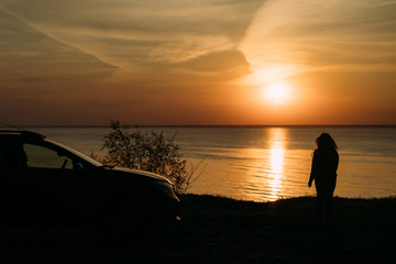 Naklejka na ściany i meble soft focus girl stands next to her car at dawn. The view from the back. Silhouette. The sun rises and the reflection falls on water. Loneliness. Privacy. The balance is inside. Good one with oneself.