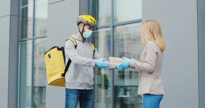 Caucasian young male courier in casque and medical mask bringing pizza to woman on street and handing to her. Deliveryman bicyclist with bag coming to customer and giving boxes.