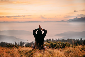  woman sits on top of a mountain at sun rise. Mountains in the fog. Warm light. The view from the back. Yoga, arms are gathered from above. Meditation. The balance of life. Appeasement. Loneliness