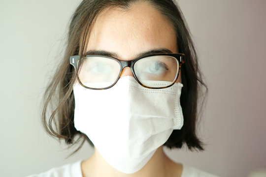 Close up of a caucasian woman wearing a white face mask during coronavirus outbreak with her glasses foggy from breath; mask problems